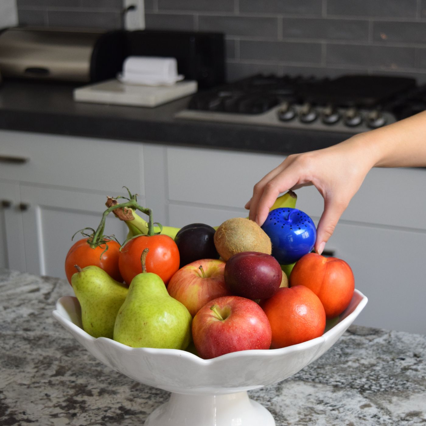 Hand placing a Carbon Bluapple in a fruit bowl with fruits and vegetables